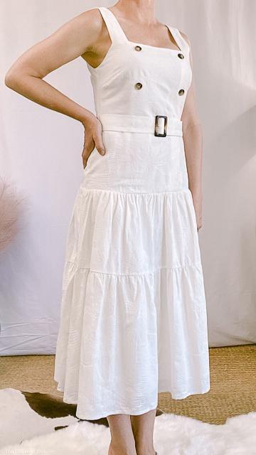 Woman wearing white sleeveless belted dress with button details, standing indoors on rug