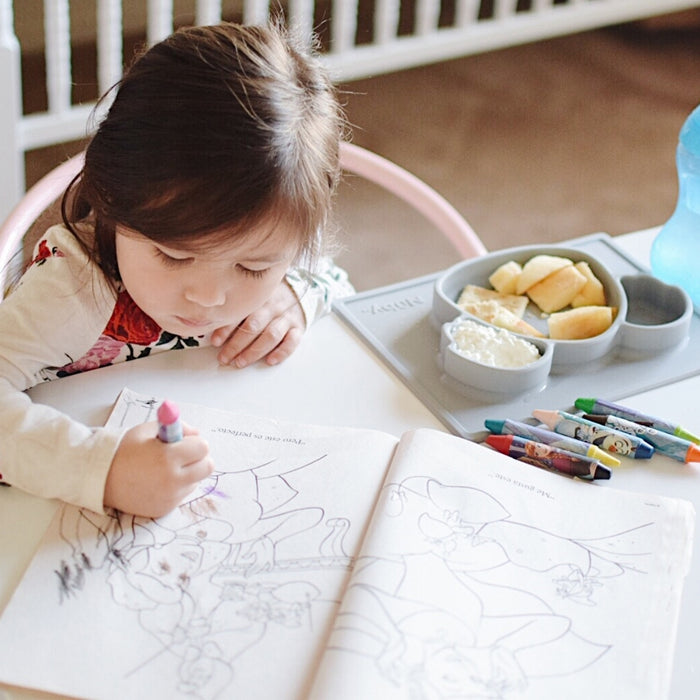 A young child colors with crayons at a small table. Snacks are served on the Nuby Sure Grip Miracle Mat All-in-One Feeding Placemat, and a blue sippy cup sits nearby as she focuses on her coloring.