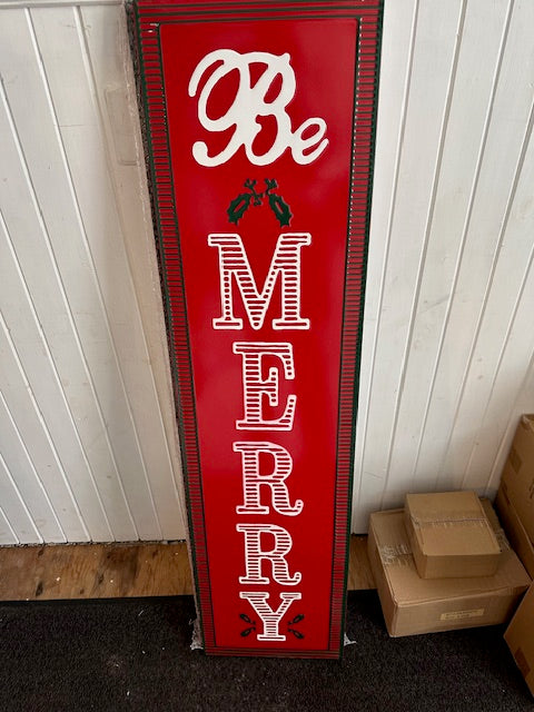 A vertical 'Be Merry' wall art decoration with a red background, white text, and Christmas-themed images including holly.