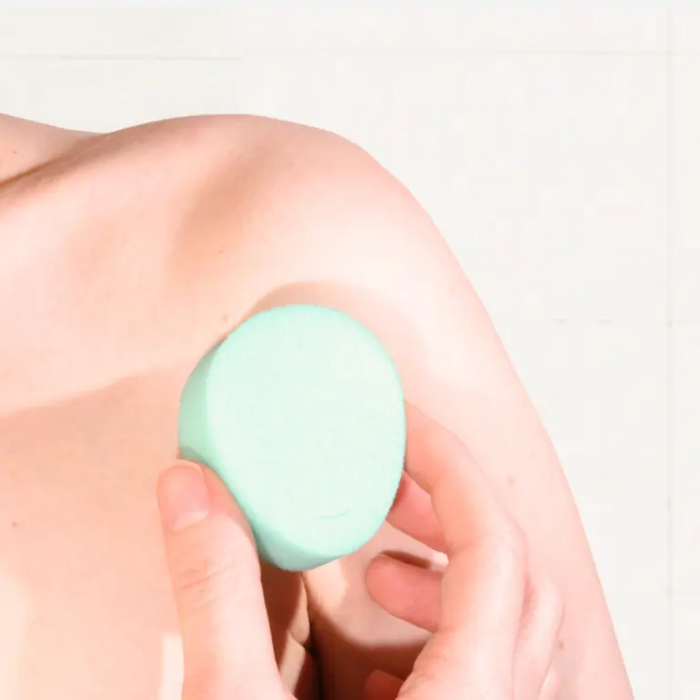 A lifestyle photograph showing a close-up of a person's hand holding a mint green sponge tampon against their upper thigh. The lighting is bright and clean, emphasizing the soft texture of the sponge against the skin.