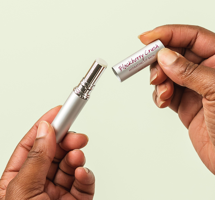 Hands holding an open Blackberry Crush Hydrating Lip Balm | Farmhouse Fresh with a sleek silver tube against a neutral background.