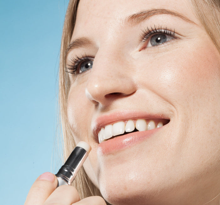 Woman smiling while applying Blackberry Crush Hydrating Lip Balm | Farmhouse Fresh to her lips against a blue background, showcasing skincare and body care wellness.