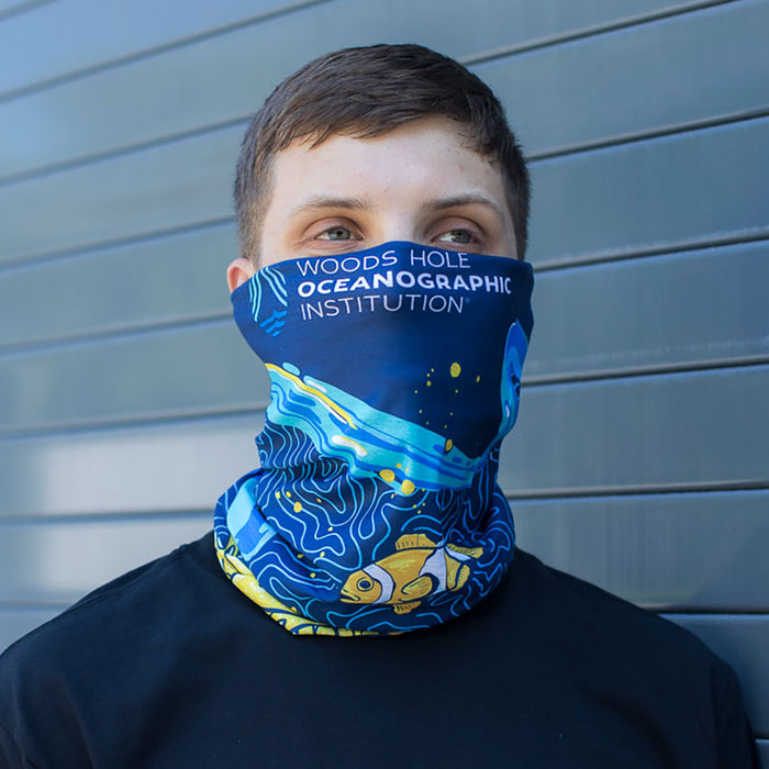 A man wearing a neck gaiter to protect himself from the sun featuring a coral reef graphic with oranges, blues and yellows.  Text says, "Woods Hole Oceanographic Institution."