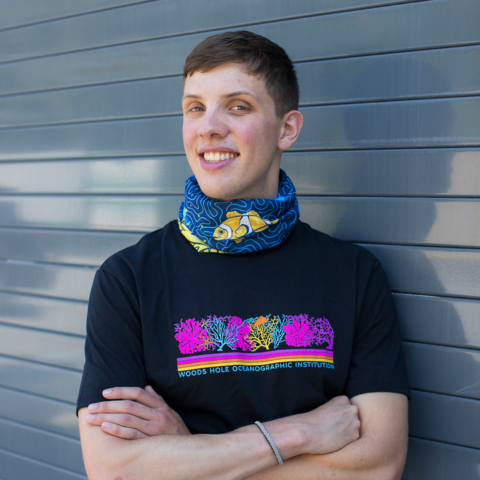 A smiling man photographed from the front wearing a neck gaiter featuring blues, oranges and yellows with a featured clownfish.