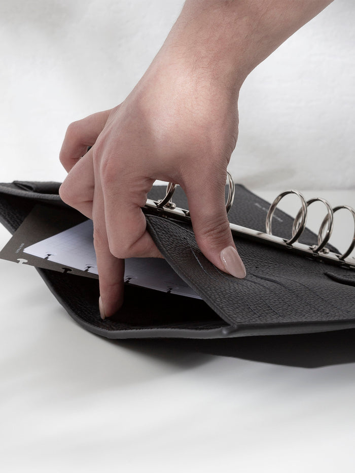 A fair-skinned hand shown opening the back pocket of the black leather agenda displayed against a shadowed background. There are inserts and a dashboard inside the pocket.