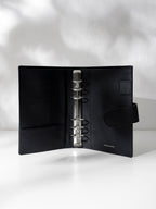 Black leather agenda displayed against a shadowed background. It is opened to show off the silver hardware and company logo stamp.