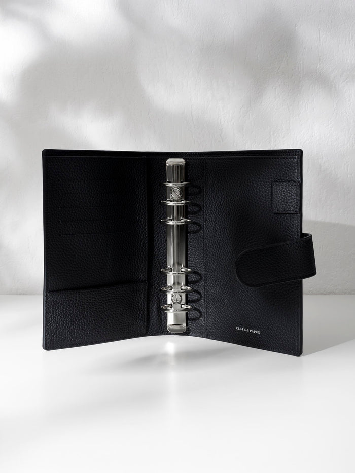 Black leather agenda displayed against a shadowed background. It is opened to show off the silver hardware and company logo stamp.
