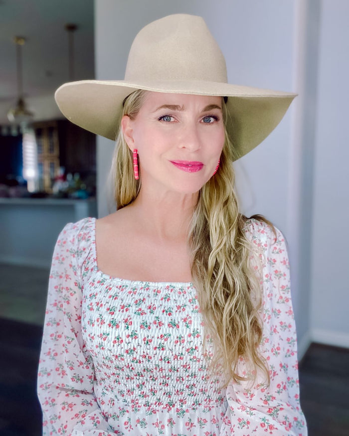 Blonde woman in a floral smocked blouse and wide beige hat, wearing pink earrings and lipstick, indoor boutique style