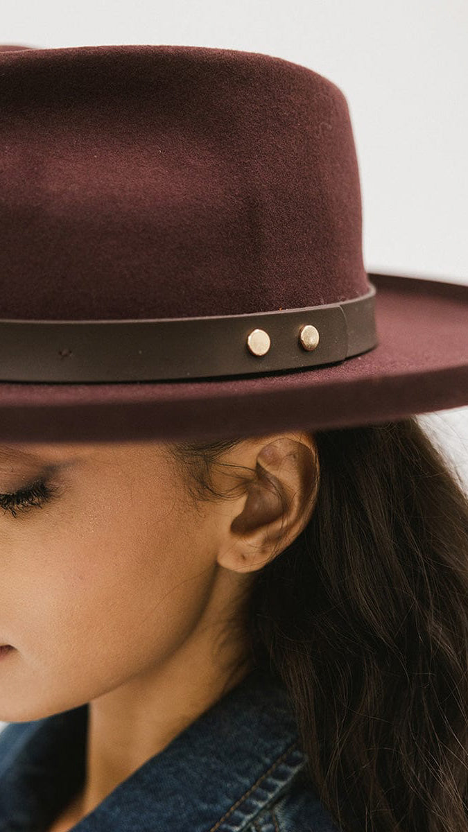 Close-up of a woman wearing a dark cherry felt hat with a brown leather band and gold studs, styled with long dark hair and denim jacket
