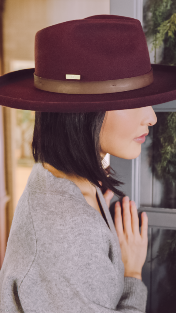 Woman wearing a stylish burgundy fedora with leather band and gray sweater indoors