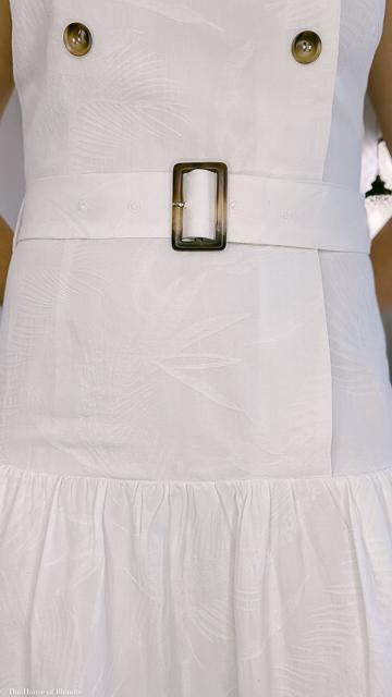 Close-up of a white sleeveless dress with tropical embossed pattern, belted waist with rectangular buckle, and button details