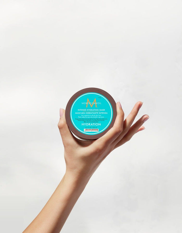 Hand holding a small jar of Intense Hydrating Mask | Moroccanoil with a turquoise label, promoting hydration and skincare benefits.