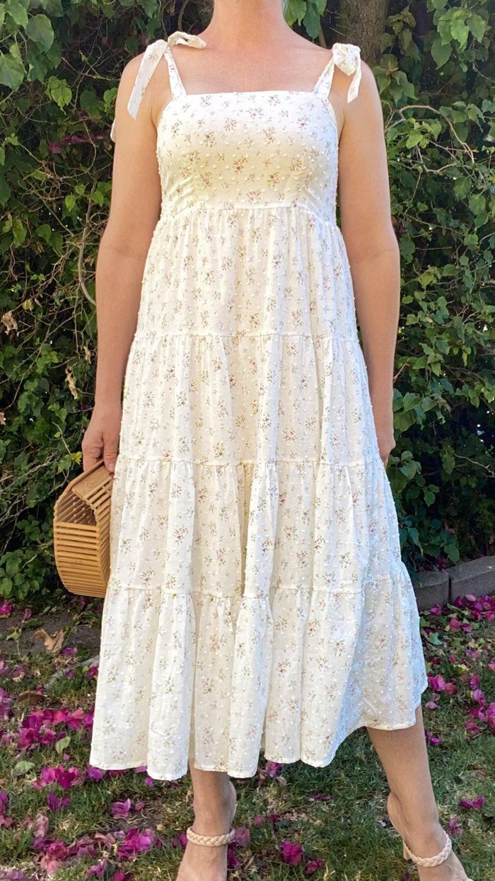 Woman wearing cream floral tiered midi dress with self-tie shoulder straps holding wooden handbag outdoors