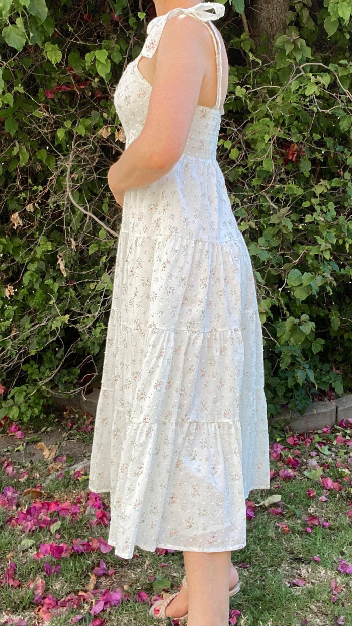 Woman wearing white floral self-tie tiered midi dress standing outdoors with green foliage and pink flowers
