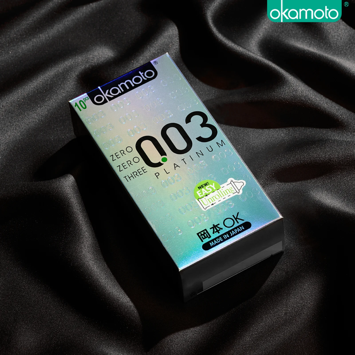 A shot of a Okamoto 003 Platinum 10-piece box resting on luxurious black silk fabric. The box features a holographic silver finish with the signature '003' logo. A bright green sticker on the front highlights the "New! Easy Unrolling" feature.
