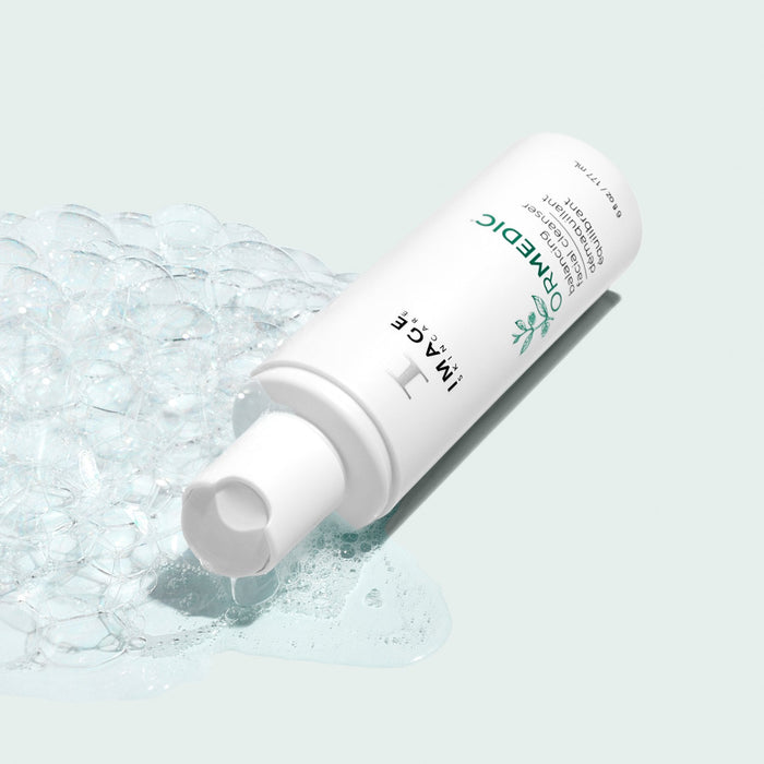 ORMEDIC® balancing facial cleanser | IMAGE Skincare bottle lying on its side with clear foam bubbles and a drop of cleanser on a light background, showcasing skincare and wellness.