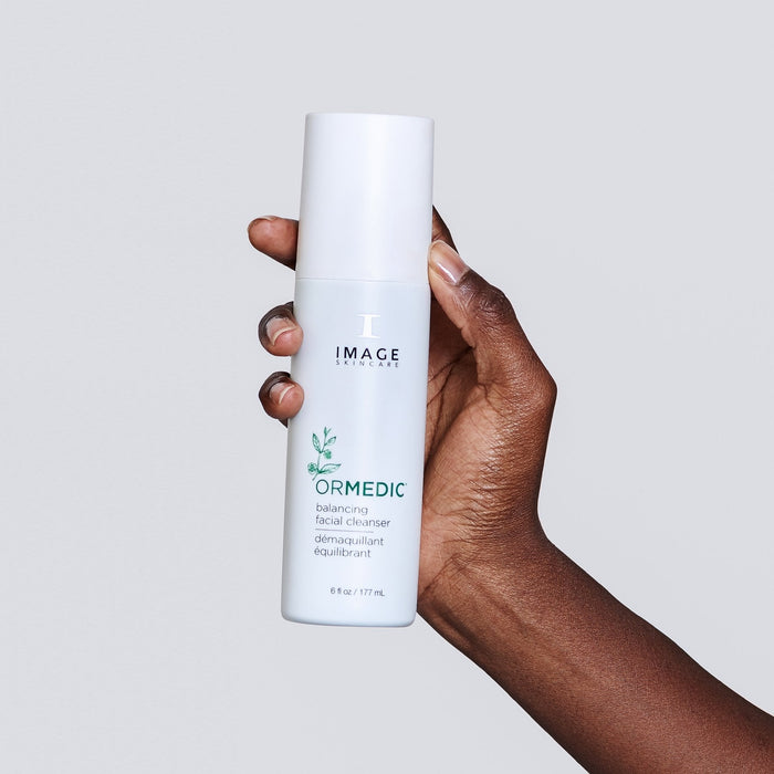 Hand holding a white bottle of ORMEDIC® balancing facial cleanser | IMAGE Skincare against a plain light gray background, showcasing the product for skincare and wellness.