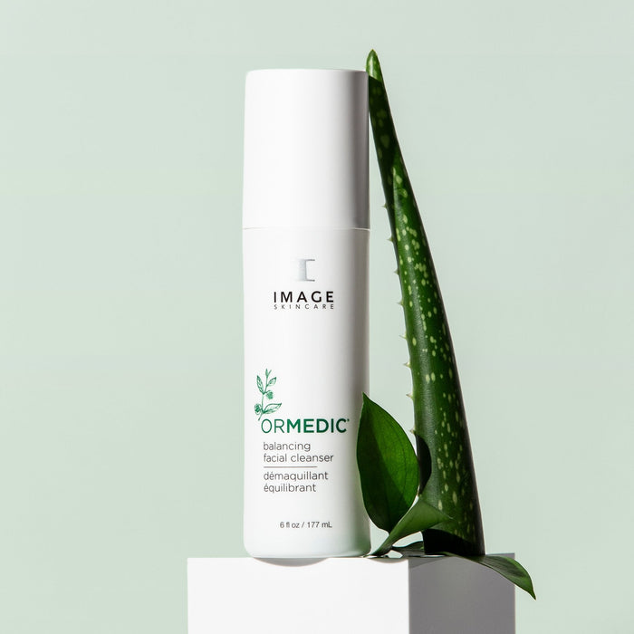 A bottle of ORMEDIC® balancing facial cleanser | IMAGE Skincare positioned next to fresh green aloe vera and leaves on a pale green background, emphasizing natural skincare and wellness.