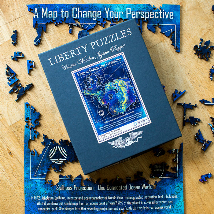A square blue box for 'Liberty Puzzles' wooden jigsaw puzzles, featuring a colorful Spilhaus Projection map on the front cover, photographed on a wooden surface with puzzle pieces scattered around the edges. The title reads, "A Map to Change Your Perspective."