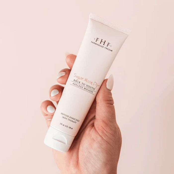 Hand holding a tube of Sugar Moon Dip® Hands | Farmhouse Fresh peptide-enriched skin therapy mousse against a soft pink background, showcasing skincare and body care product.