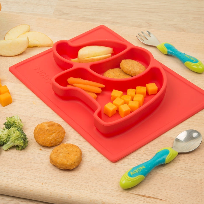A Nuby Sure Grip Miracle Mat All-in-One Feeding Placemat, shaped like a red dinosaur, holds apple slices, cheese cubes, carrot sticks, and chicken nuggets. A colorful baby spoon and fork rest on the wooden table nearby.