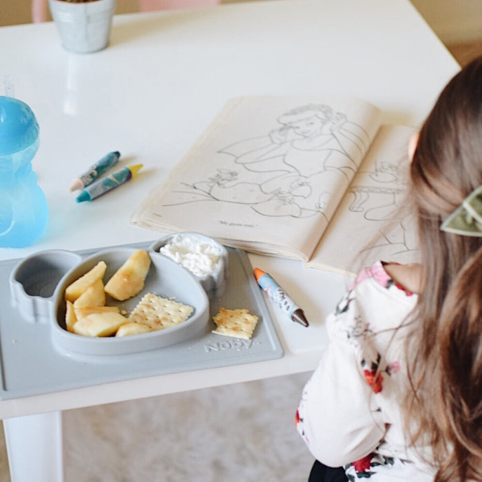 A young child with long brown hair colors at a white table, enjoying snacks served on the Nuby Sure Grip Miracle Mat All-in-One Feeding Placemat, which keeps apple slices, crackers, and dip secure beside her blue sippy cup and crayons.