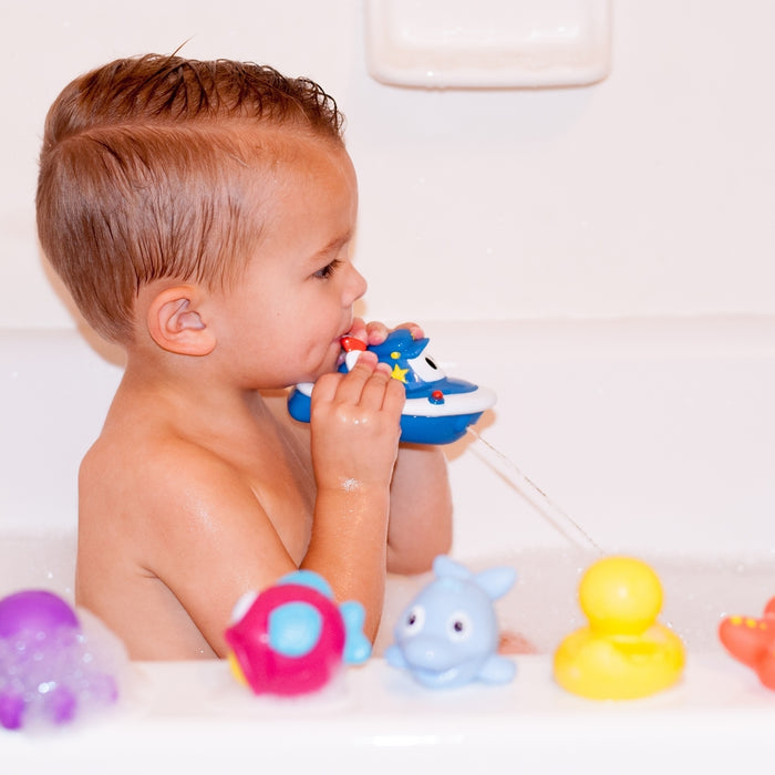 A young child with short, wet hair enjoys a bubble bath, playing with a Nuby Tub Tugs (2 Pack) boat that sprays water. Colorful floating toys, like a duck and dolphin, surround them for playful bath time fun.
