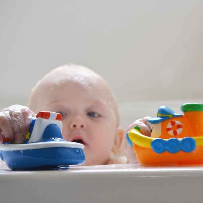 A baby in a bathtub plays with Nuby Tub Tugs (2 Pack) colorful floating boat toys, focusing intently while only their head and hands appear above the tub’s edge.