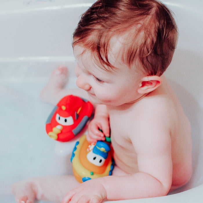A baby with wet hair relaxes in the tub, playing with Nuby's Tub Tugs (2 Pack)—colorful red and yellow bath boats with cartoon faces—enjoying the water and floating toys.