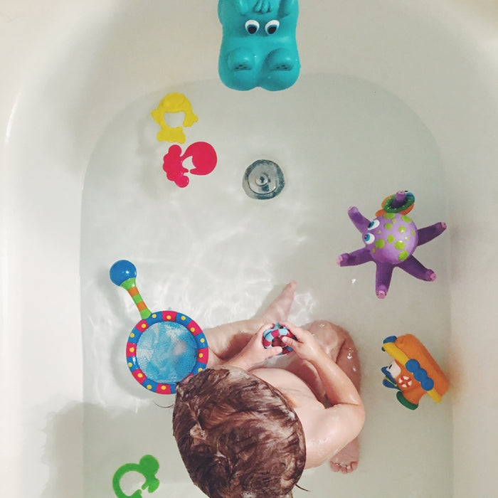A toddler plays in the bath with Nuby Tub Tugs (2 Pack), surrounded by colorful toys like a blue hippo, purple octopus, floating animal shapes, and baby bath boats, while holding a small toy in both hands.