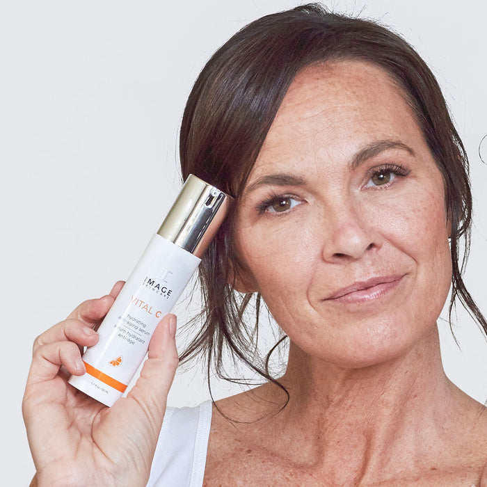 Woman with clear skin holding a bottle of VITAL C hydrating anti-aging serum | IMAGE Skincare, showcasing skincare product for hydration and anti-aging benefits.