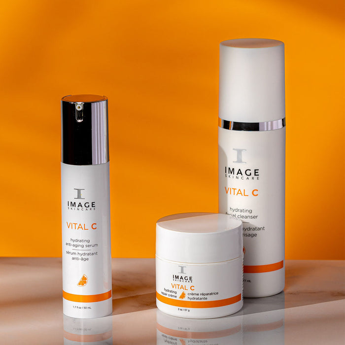 Three IMAGE Skincare products from the VITAL C line, including the VITAL C hydrating anti-aging serum | IMAGE Skincare, displayed on a reflective surface with an orange background.