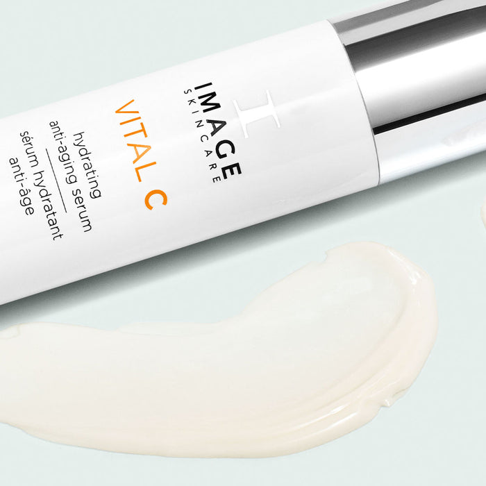 Close-up of a white and silver bottle of VITAL C hydrating anti-aging serum | IMAGE Skincare next to a smooth, creamy serum texture on a light background.
