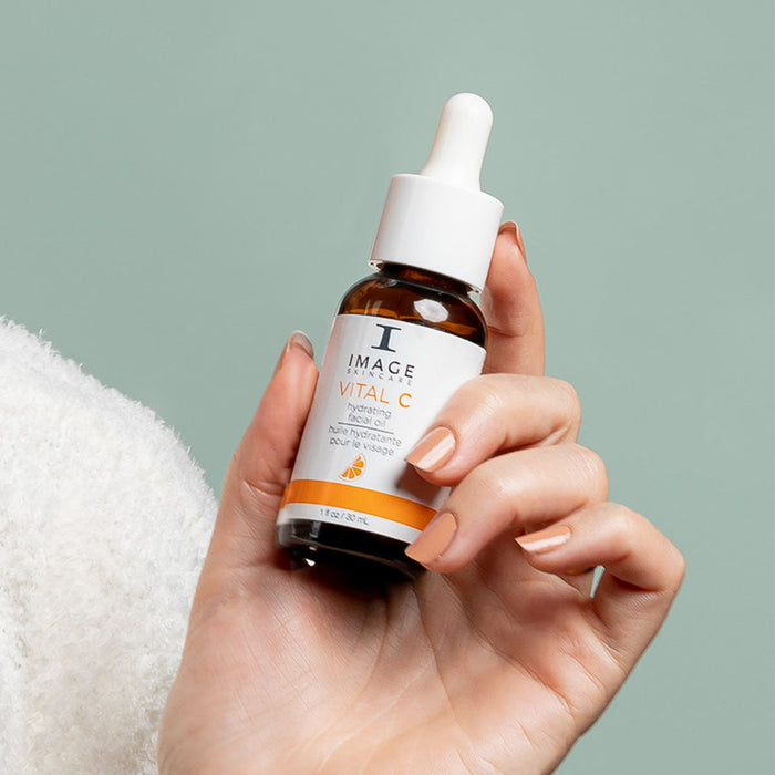 Hand holding a brown glass dropper bottle of VITAL C hydrating facial oil | IMAGE Skincare against a soft green background, showcasing skincare product for hydration and wellness.