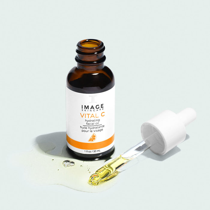 Open amber glass bottle of VITAL C hydrating facial oil | IMAGE Skincare with dropper releasing light yellow oil on a pale background for skincare and wellness.